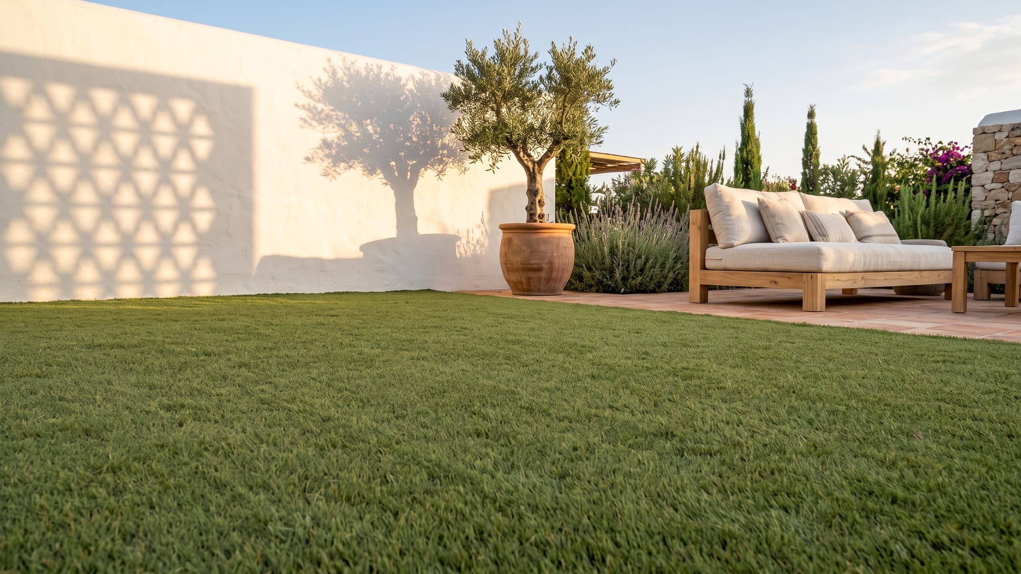 Jardín mediterráneo al atardecer con una superficie amplia de césped artificial, muro encalado con la sombra de una celosía, un olivo en tinaja de barro y una zona de estar baja de madera clara